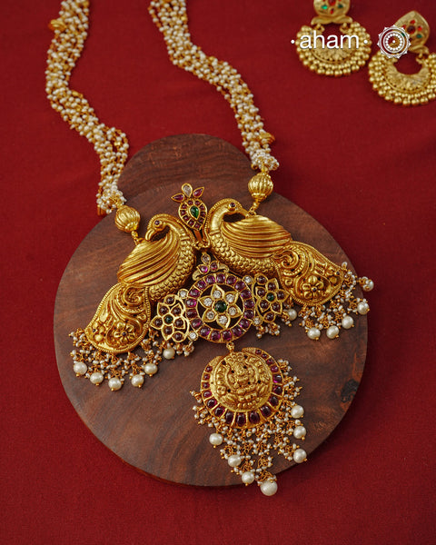 Crafted in 92.5 sterling silver with a rich fine&nbsp;gold polish, this statement neckpiece is a celebration of timeless Indian artistry. At its heart, a pair of majestic peacocks gracefully frame an intricately detailed pendant, adorned with vibrant ruby and emerald-toned stones, sparkling kundan work, and a finely sculpted Lakshmi motif. Delicate freshwater pearl clusters lend graceful movement, while the hand-strung pearl chain completes the design with understated elegance.