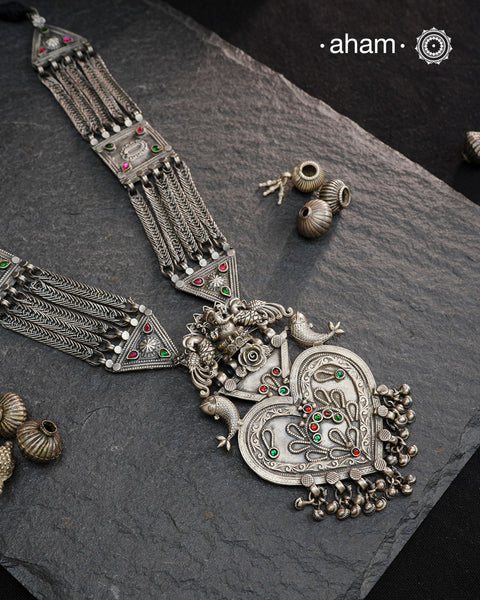 This Tribal Silver Neckpiece artfully merges vintage and modern design elements. The centerpiece pendant features a heart-shaped amulet crowned by Ganeshji, with intricately detailed peacocks and fishes on either side. The neckpiece is completed with a classic Jaisalmeri-style silver belt, showcasing exceptional traditional craftsmanship. Perfect for those who value heritage artistry in their jewelry collection.