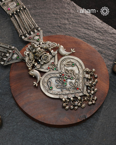 This Tribal Silver Neckpiece artfully merges vintage and modern design elements. The centerpiece pendant features a heart-shaped amulet crowned by Ganeshji, with intricately detailed peacocks and fishes on either side. The neckpiece is completed with a classic Jaisalmeri-style silver belt, showcasing exceptional traditional craftsmanship. Perfect for those who value heritage artistry in their jewelry collection.