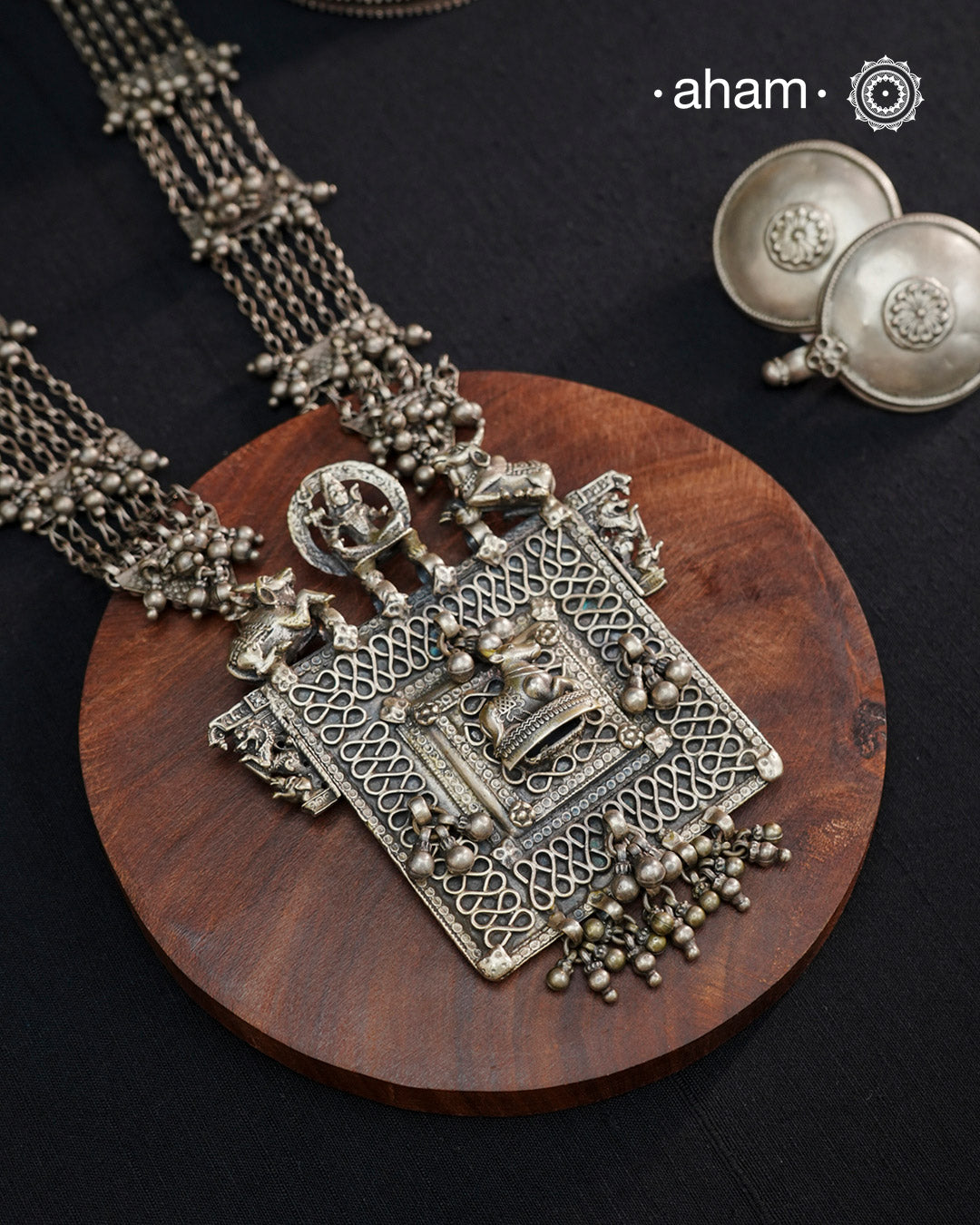 This expertly crafted silver neckpiece features a square amulet intricately depicting Lord Shiva alongside Nandi, embodying traditional tribal artistry. The side chains adorned with numerous ghungroos add a rhythmic charm, enhancing its cultural authenticity. Perfect for those who appreciate heritage craftsmanship and spiritual symbolism in their jewelry collection.