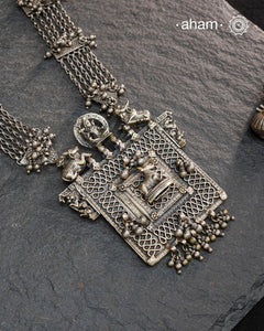 This expertly crafted silver neckpiece features a square amulet intricately depicting Lord Shiva alongside Nandi, embodying traditional tribal artistry. The side chains adorned with numerous ghungroos add a rhythmic charm, enhancing its cultural authenticity. Perfect for those who appreciate heritage craftsmanship and spiritual symbolism in their jewelry collection.