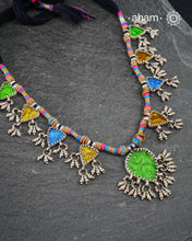This Tribal Silver Neckpiece showcases a vibrant green glass pendant at its center, enhanced by meticulously hand-tied tiny ghungroos. On each side, three finely crafted silver leaves create a harmonious and elegant design. Ideal for layering during Navratri, this piece blends traditional artistry with festive sophistication to complement your celebration attire.
