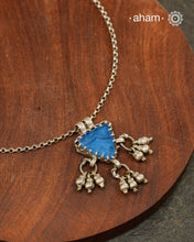 A charming piece inspired by traditional tribal adornments, this neckpiece features a vibrant blue glass pendant accented with silver ghungroos that sway gently with movement. The striking colour of the glass contrasts beautifully with the rustic silver finish, creating a look that is both distinctive and effortless.

Lightweight and easy to style, it pairs beautifully with handloom sarees, cotton kurtas, dresses, and everyday separates. Its understated size and playful detailing make it perfect for those w