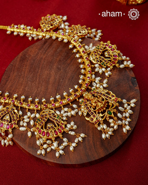 Crafted in 92.5 sterling silver with an exquisite fine gold polish, this Guttapusalu-inspired neckpiece is a beautiful expression of South Indian craftsmanship. Intricately adorned with vibrant kemp stones, shimmering kundan work, and graceful Lakshmi motif in the center, each pendant is framed by delicate clusters of freshwater pearls that lend the piece its signature elegance and movement.