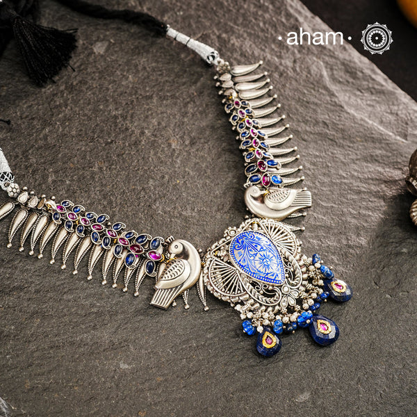 Meticulously crafted from 92.5 sterling silver, this exquisite Ira Silver Neckpiece features a central meena work pendant flanked by intricate peacock motifs. The design is enhanced with vibrant blue and maroon kemp stone accents, creating a striking contrast against the silver. Designed to rest elegantly below the neck, this piece combines traditional Indian artistry with sophisticated craftsmanship for a timeless addition to your jewelry collection.