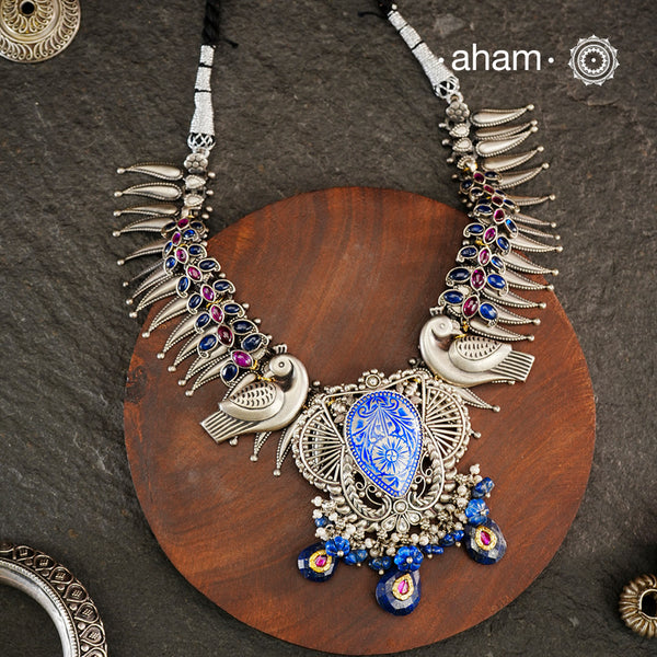 Meticulously crafted from 92.5 sterling silver, this exquisite Ira Silver Neckpiece features a central meena work pendant flanked by intricate peacock motifs. The design is enhanced with vibrant blue and maroon kemp stone accents, creating a striking contrast against the silver. Designed to rest elegantly below the neck, this piece combines traditional Indian artistry with sophisticated craftsmanship for a timeless addition to your jewelry collection.