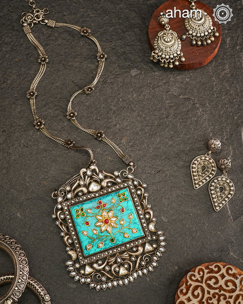 Make a bold statement with the Ira Turquoise Silver Neckpiece! This stunning piece features a vibrant turquoise stone set in silver with intricate inlay work and kundan all around it.  Delicate flowers decorate the chain, all crafted in 92.5 silver. Elevate your style and stand out from the crowd with this unique necklace.