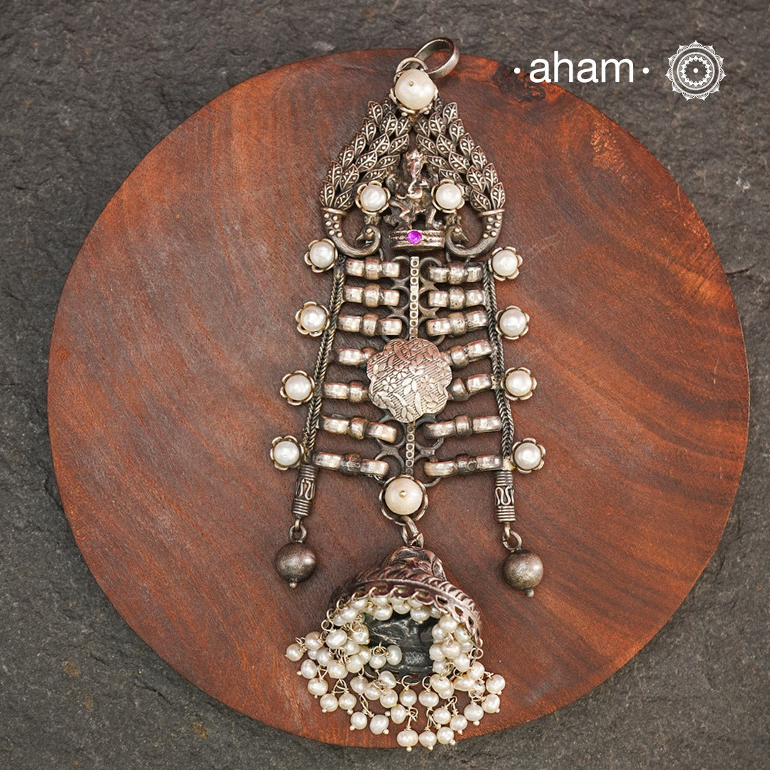 This unique Ganesha Pendant is a statement piece crafted in 92.5 silver. The intricate design features Ganesha at the top, flanked by delicately crafted peacocks on either side. A jhumka hangs at the bottom, adorned with pearl highlights for a touch of elegance. Elevate any outfit with this one-of-a-kind pendant.