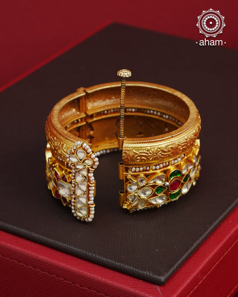 This openable Kundan Gold Polish Silver Kada gives the illusion of wearing two stacked kadas, with exquisite kundan work and laced pearls adding to its charm. Made with silver and finished in gold polish, it's a must-have for any jewellery collection.