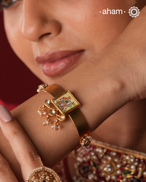 Crafted in 92.5 Sterling Silver and finished with a rich 24 Carat Gold Polish, this handcrafted cuff is a celebration of devotion, artistry, and India's enduring craft traditions. At its heart lies a miniature hand-painted portrait of Shrinathji, meticulously created by skilled artisans, making every piece truly one of a kind.
Framed within an elegant square bezel and complemented by delicate freshwater pearls and kundan-inspired detailing,the cuff draws inspiration from the revered Pichwai art of Nathdwara