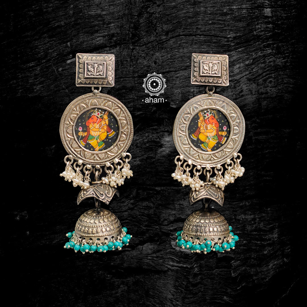 Handcrafted 92.5 sterling silver jhumkie earrings. The highlight of this piece is intricate miniature hand painted Ganesha Motif in vibrant colours enclosed with a glass top and floral motifs in the back. These are hand painted one of a kind wearable art pieces. 